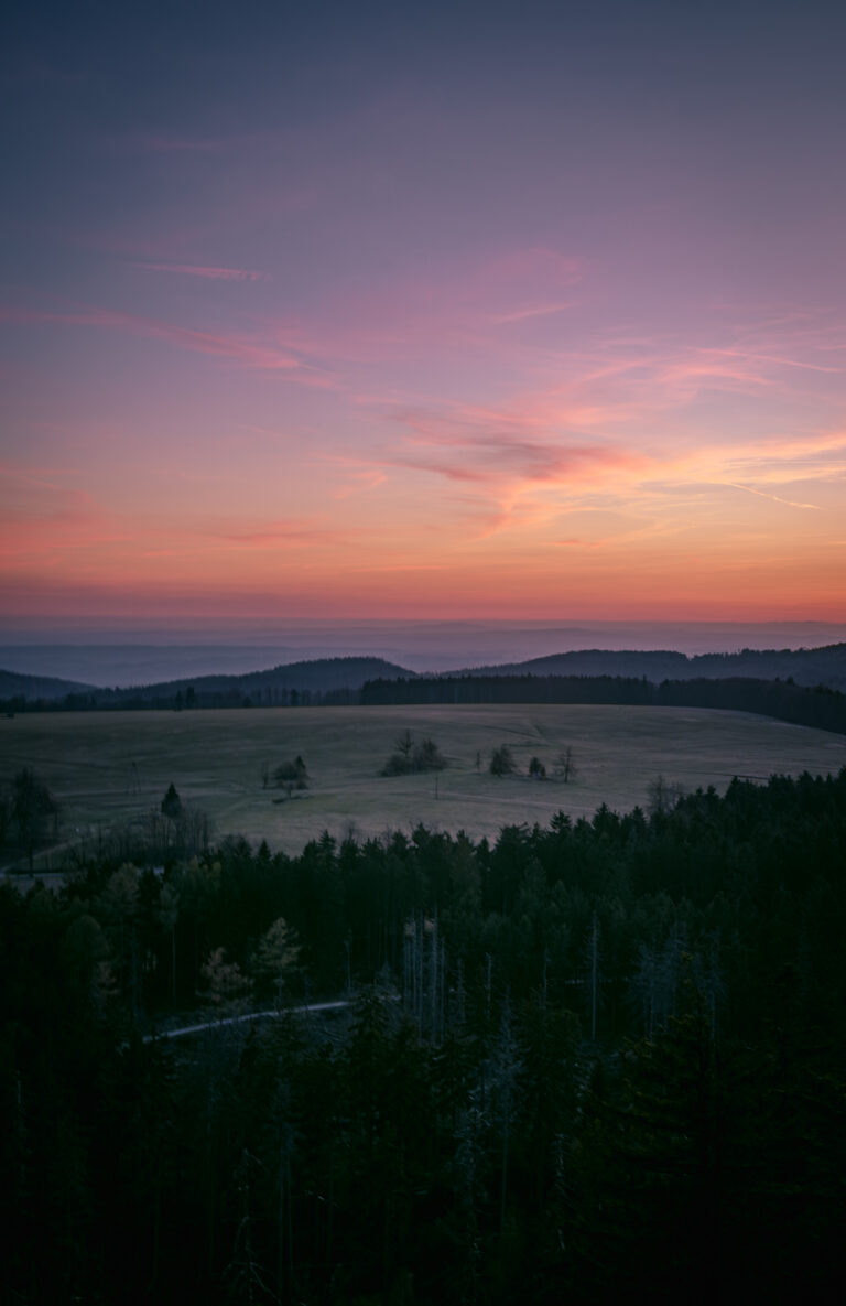 the evening sky in shades of purple and orange, along with small tree-covered mountains and a vast meadow
